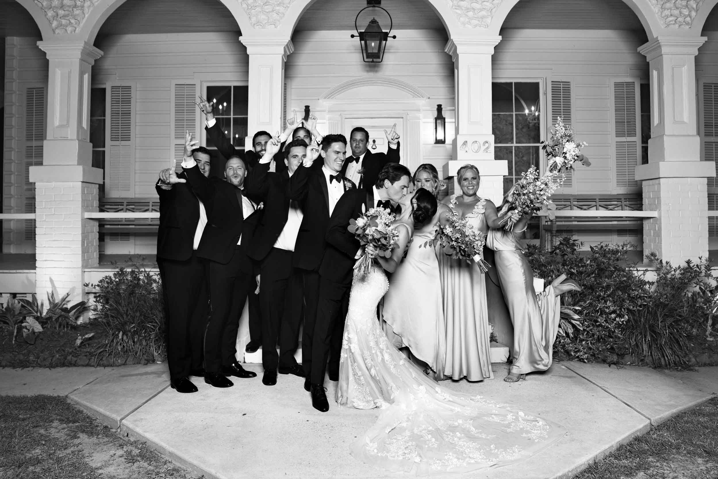 bride and groom kissing with wedding party celebrating in front of Maison Lafitte in Covington, Louisiana