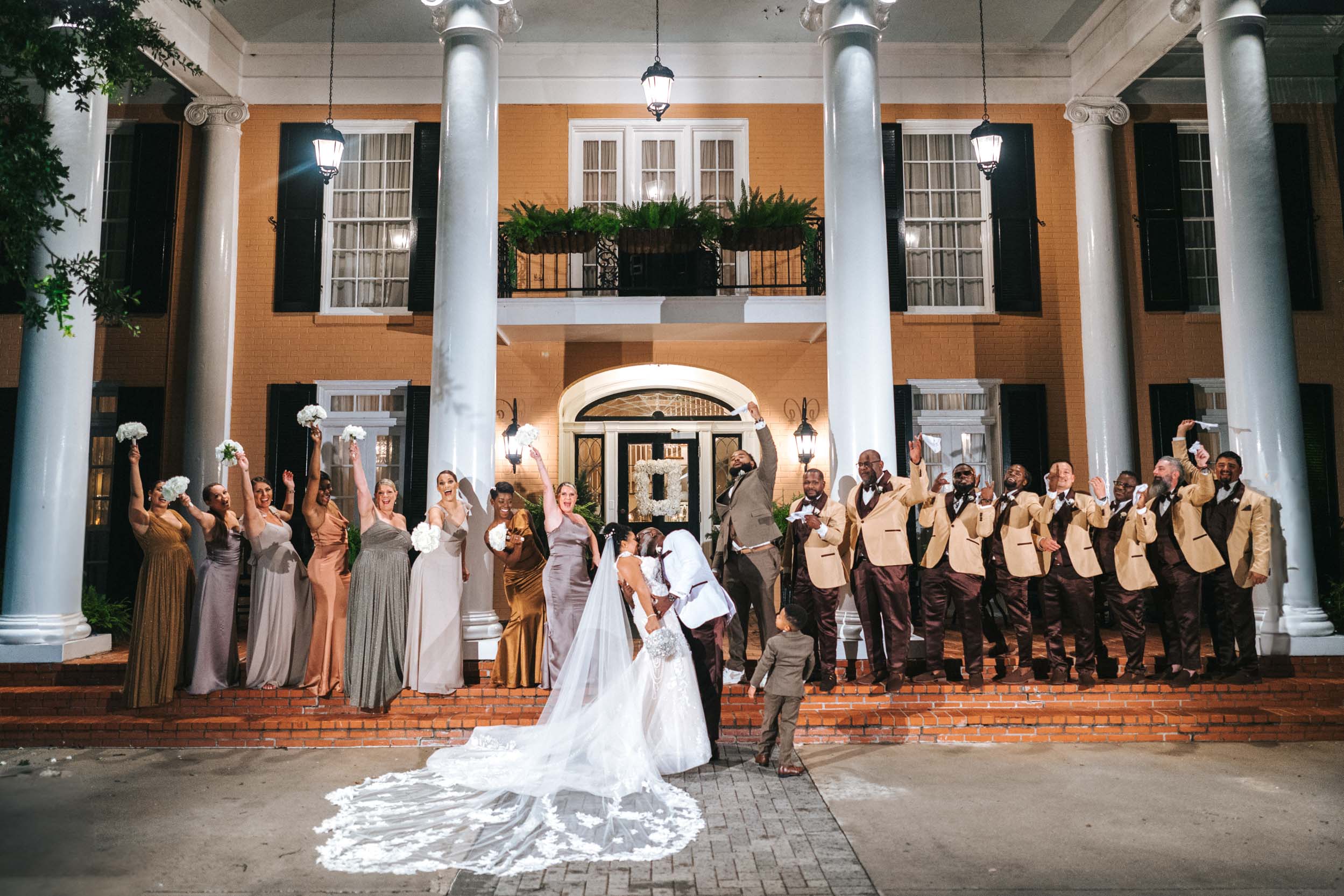 African American bride and groom kissing with wedding party celebrating on the front steps of The Southern Oaks in New Orleans