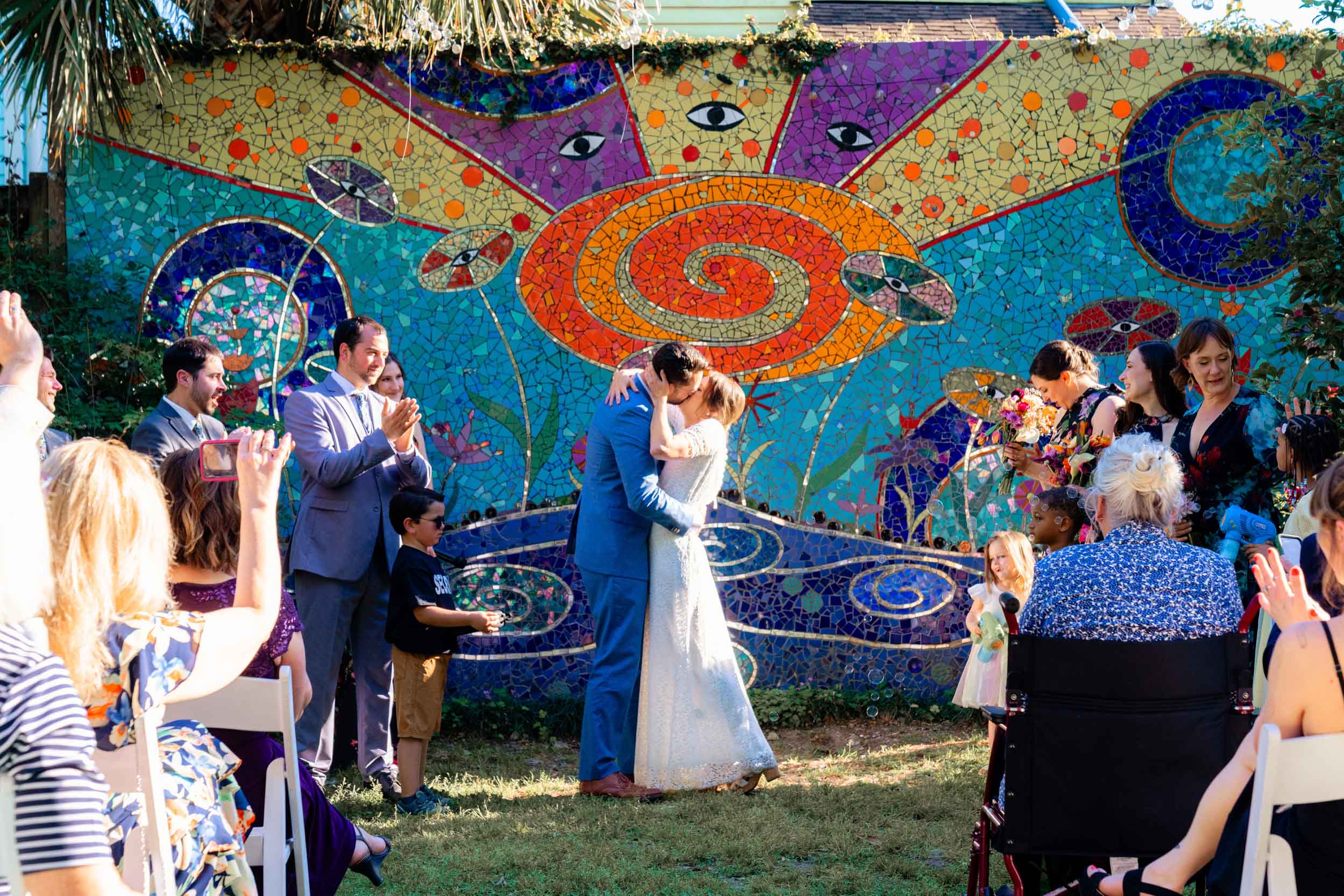 bride and groom kissing during wedding ceremony in front of colorful mural at Clouet Gardens in the Bywater of New Orleans