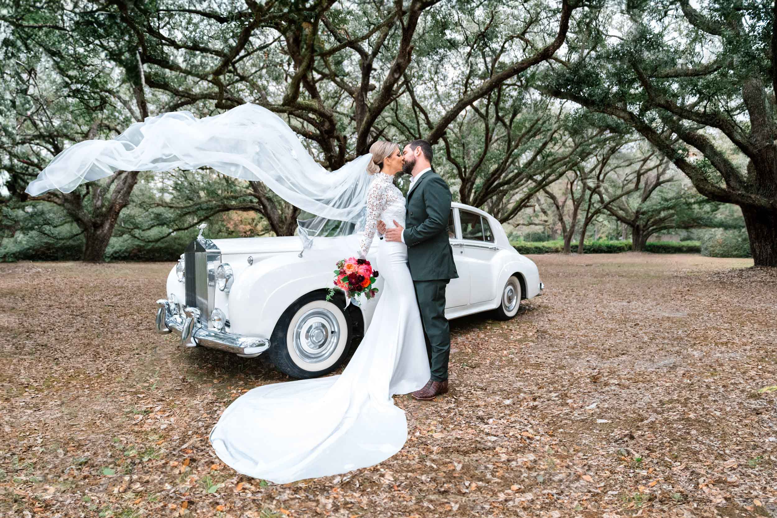 bride and groom kissing with veil flying in the air in front of Rolls-Royce and large oak trees at The Greenwood Covington, Louisiana