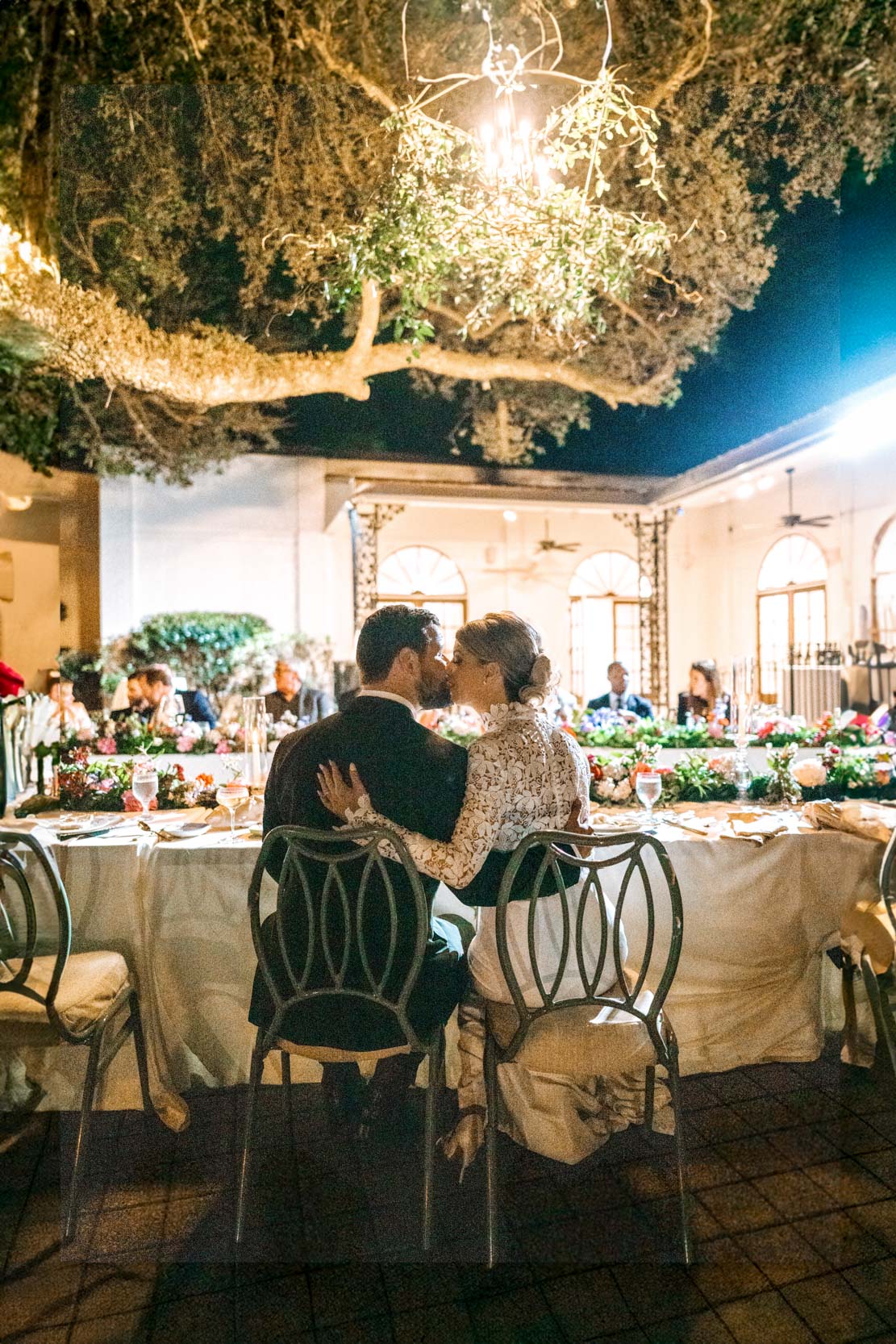 bride and groom sitting and kissing each other during wedding reception under outdoor chandelier and oak tree at The Greenwood Covington, Louisiana