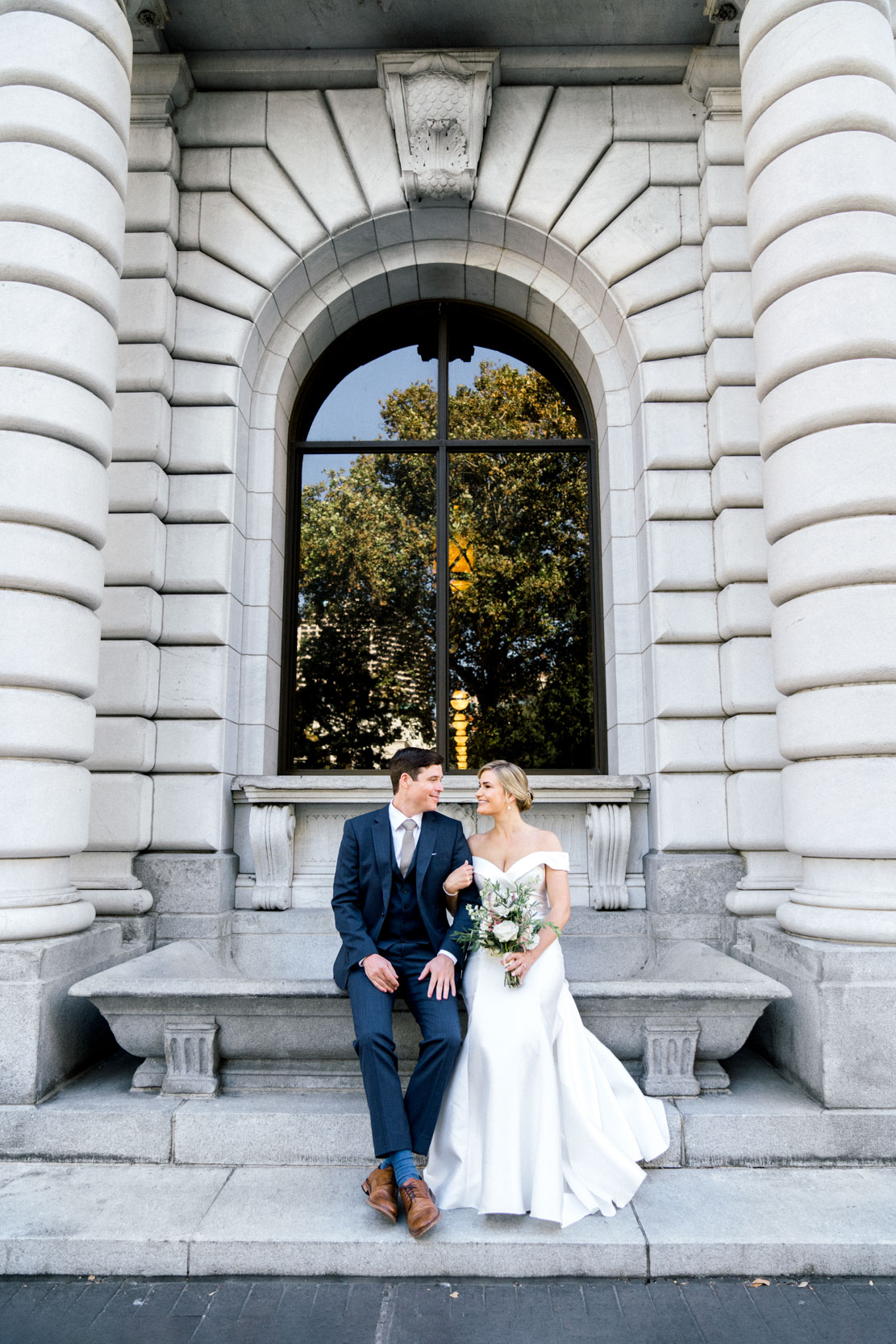 bride and groom sitting on bench in front of strone masonry building, John Minor Wisdom Court of Appeals in Lafayette Square Park in New Orleans