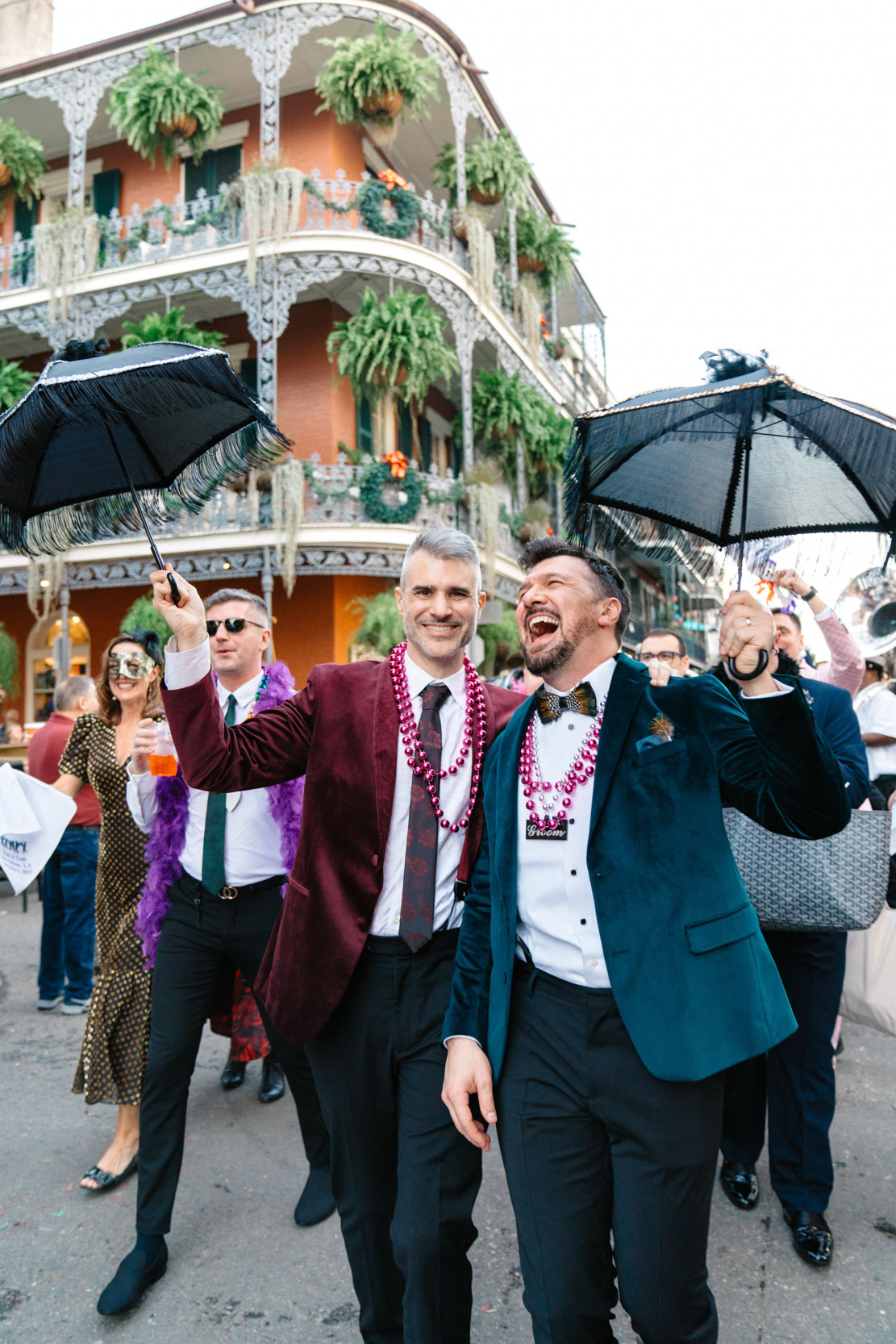 gay couple celebrating during their 2nd line band procession on wedding day through the New Orleans French Quarter ith ferns and beautiful mansion in the background