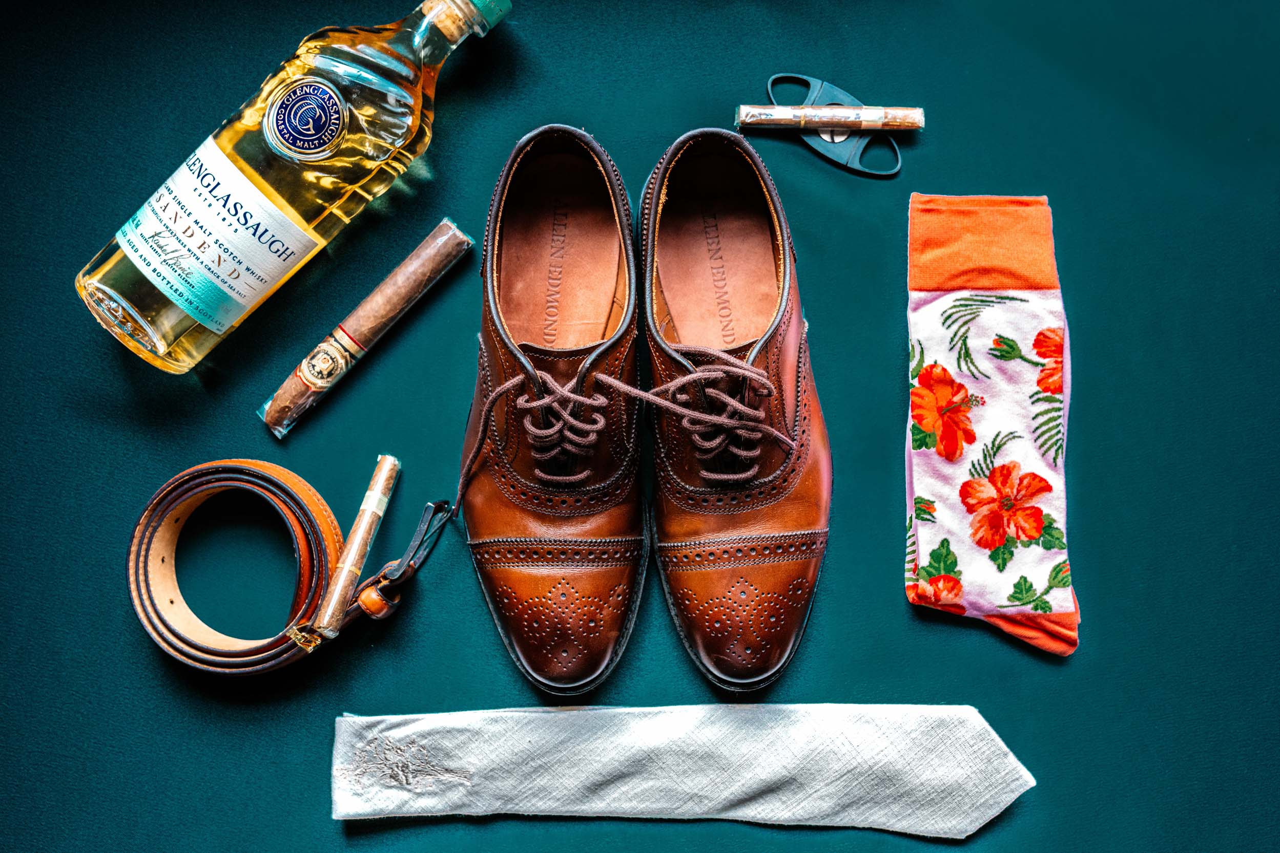 green flat lay of groom's leather shoes, belt, cigar, bottle of Glenglassaugh scotch, floral socks, and ivory tie at Margaret Place in New Orleans