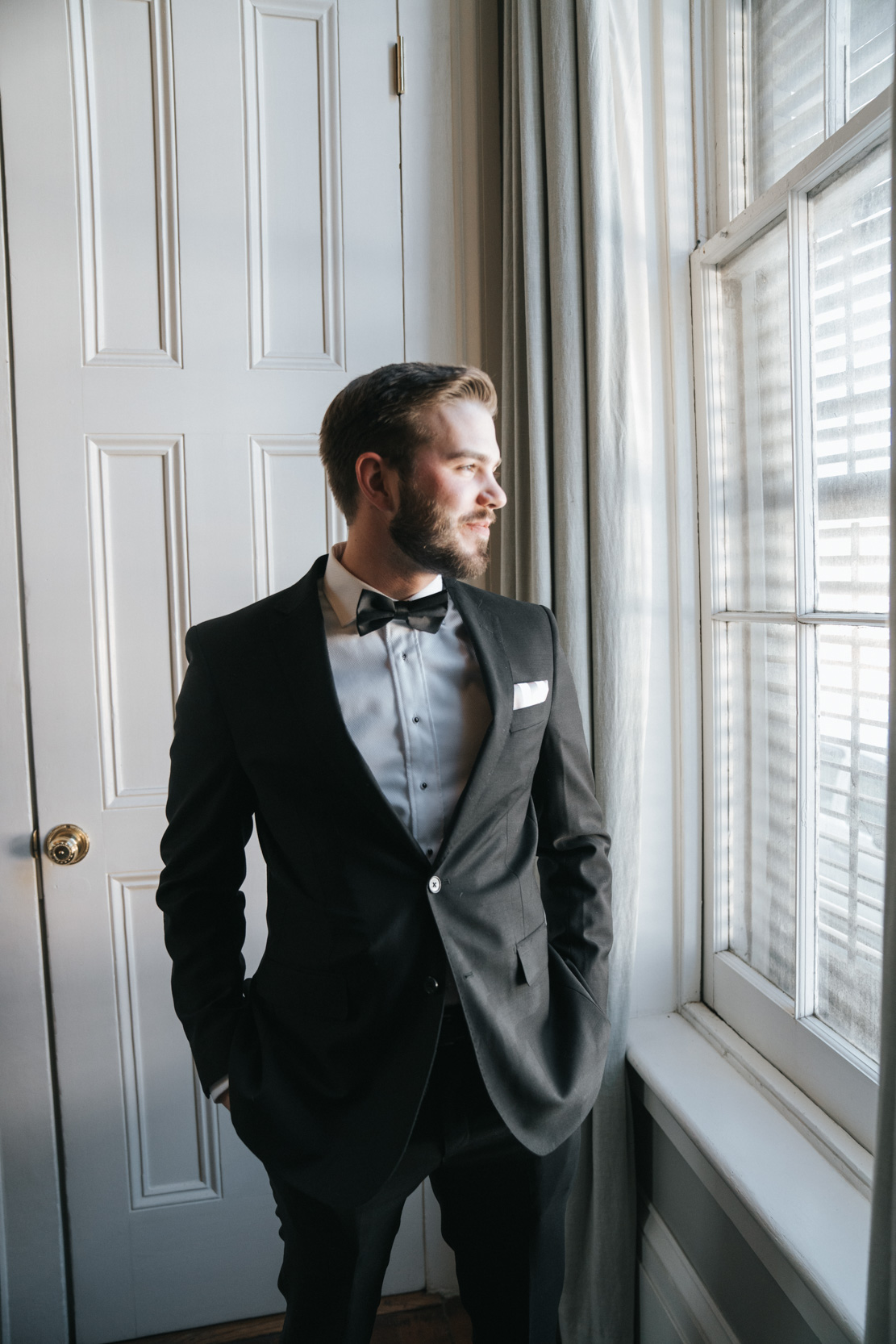 groom in black suit smiling and looking out of window on wedding day at the Terrell House in New Orleans