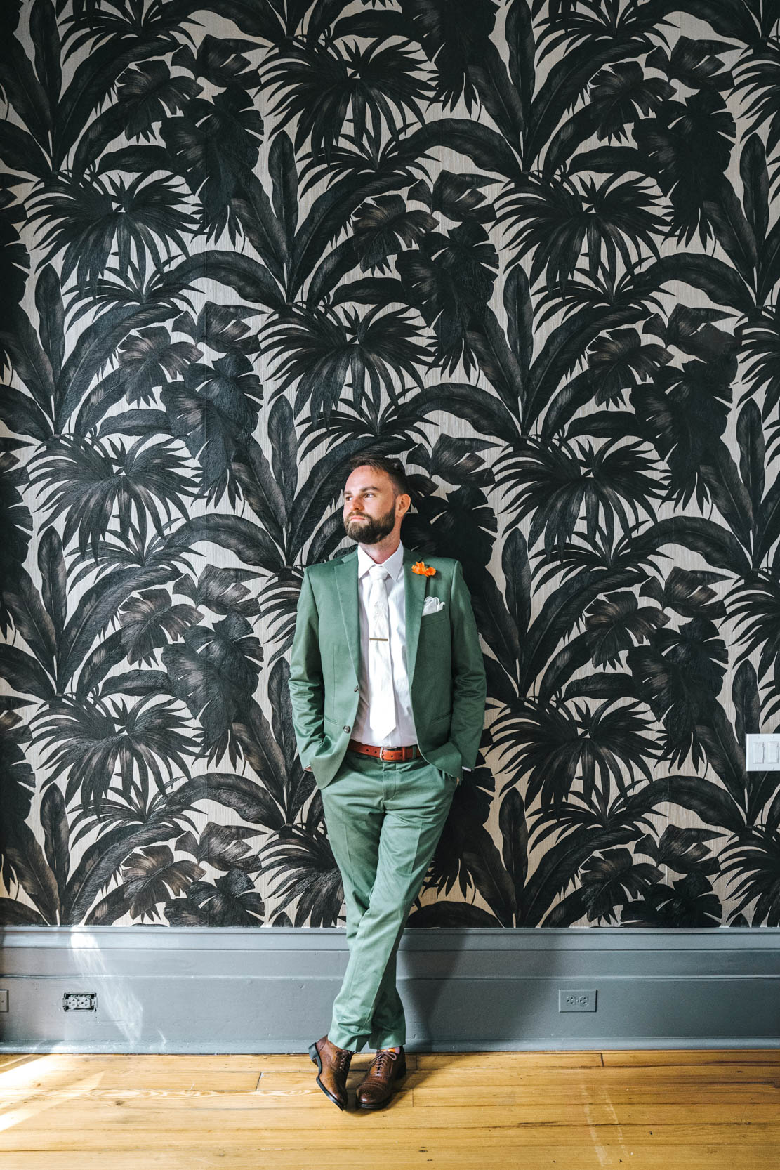 groom posing in green suit in front of floral wall design on wedding day at Margaret Place in New Orleans