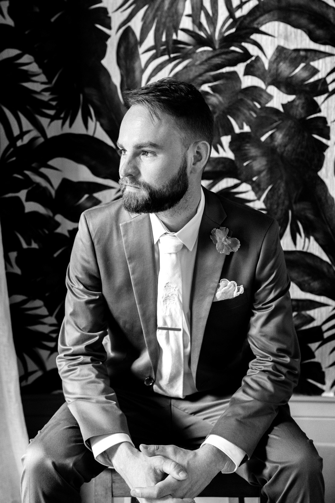 groom posing in green suite on chair in front of elaborate floral wall design at Margaret Place in New Orleans