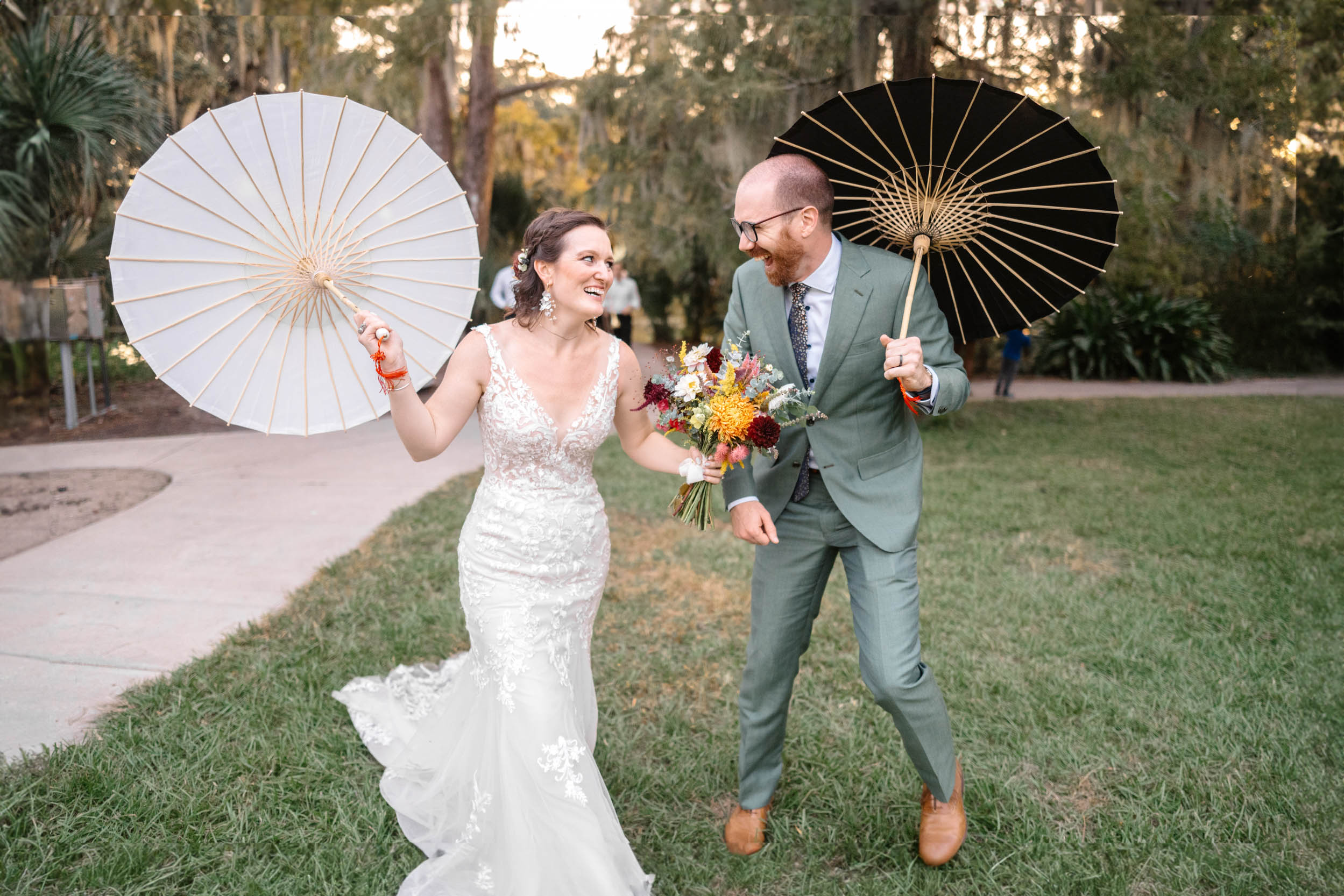 young couple laughing and holding parasols during 2nd line band procession and Baby t-rex farms flowers on wedding day in New Orleans City Park