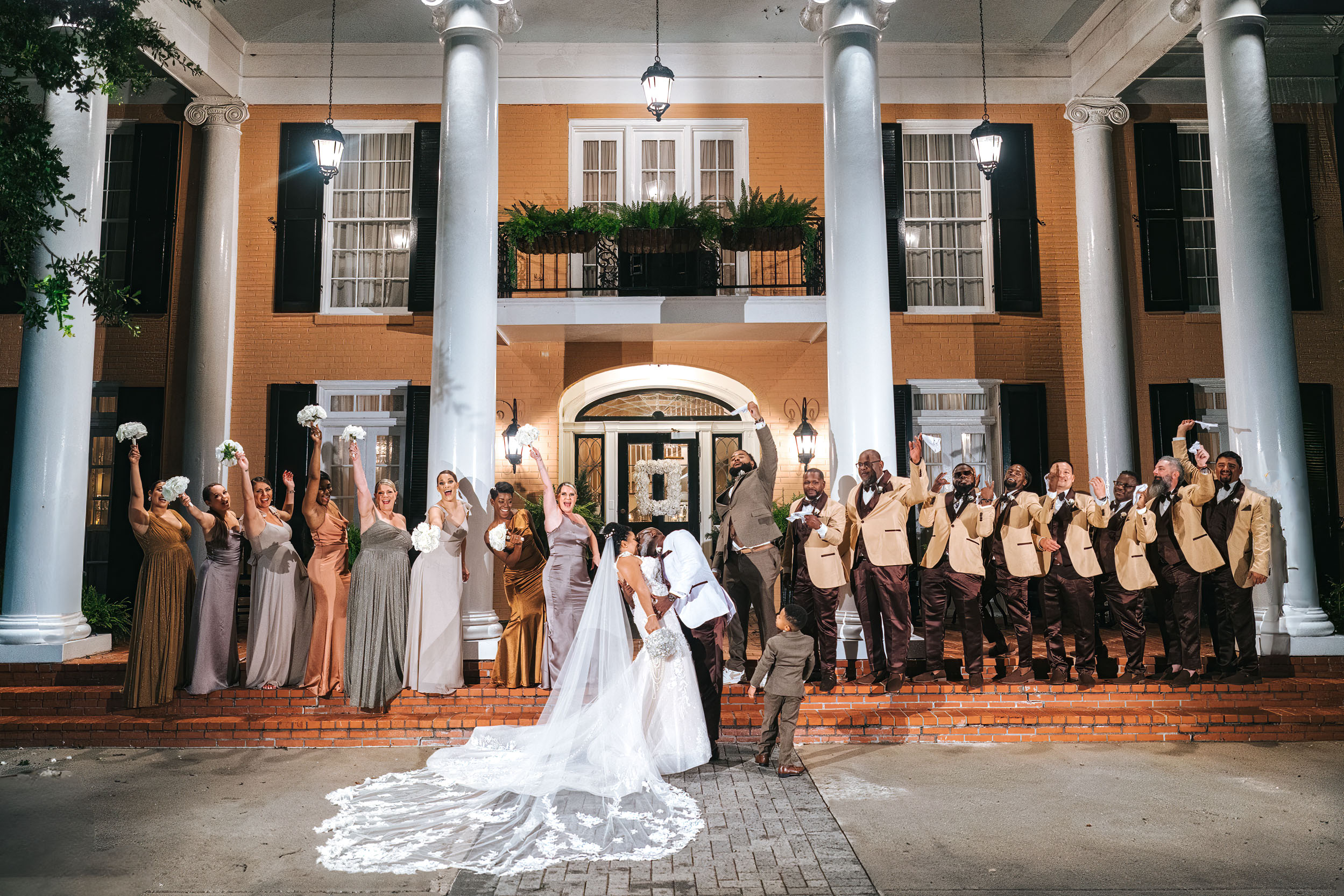 African American bride and groom kissing with wedding party celebrating on the front steps of The Southern Oaks in New Orleans
